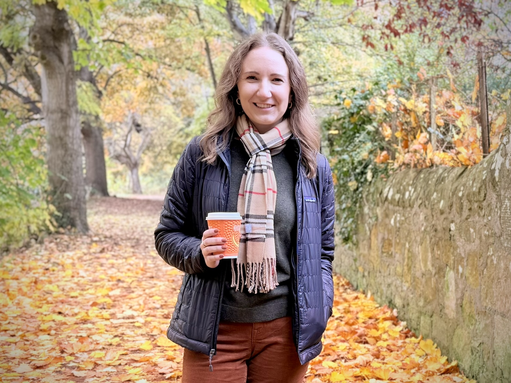 A mom holding a coffee cup on a winding path with fall leaves behind her